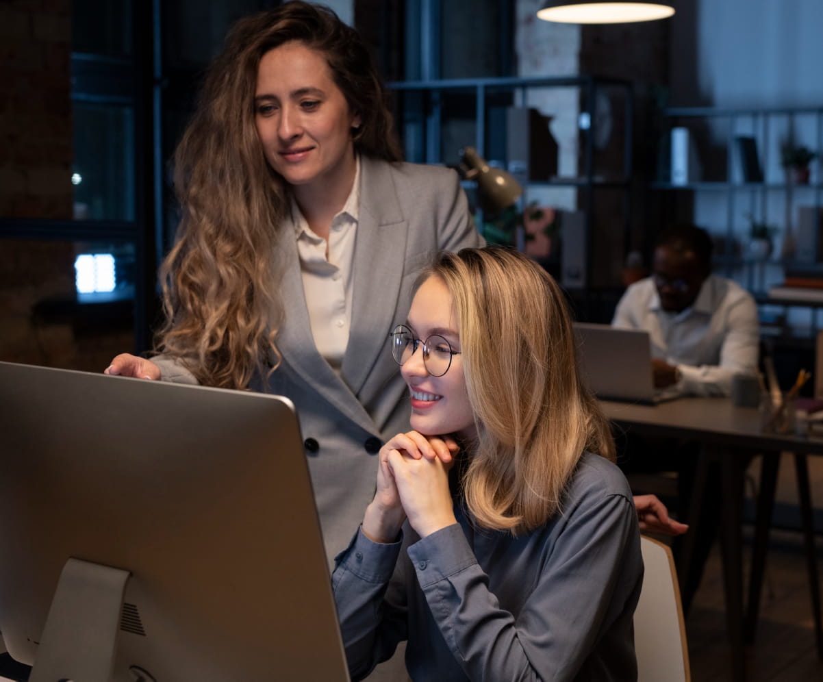 A workstation with few people and computers, two girls discussing each other about their tasks in front of a computer